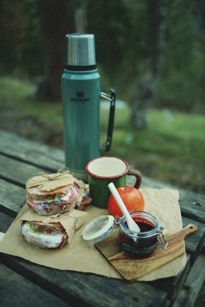 Cozy outdoor breakfast setup featuring sandwiches, thermos, and fruit on a rustic picnic table in a forest.